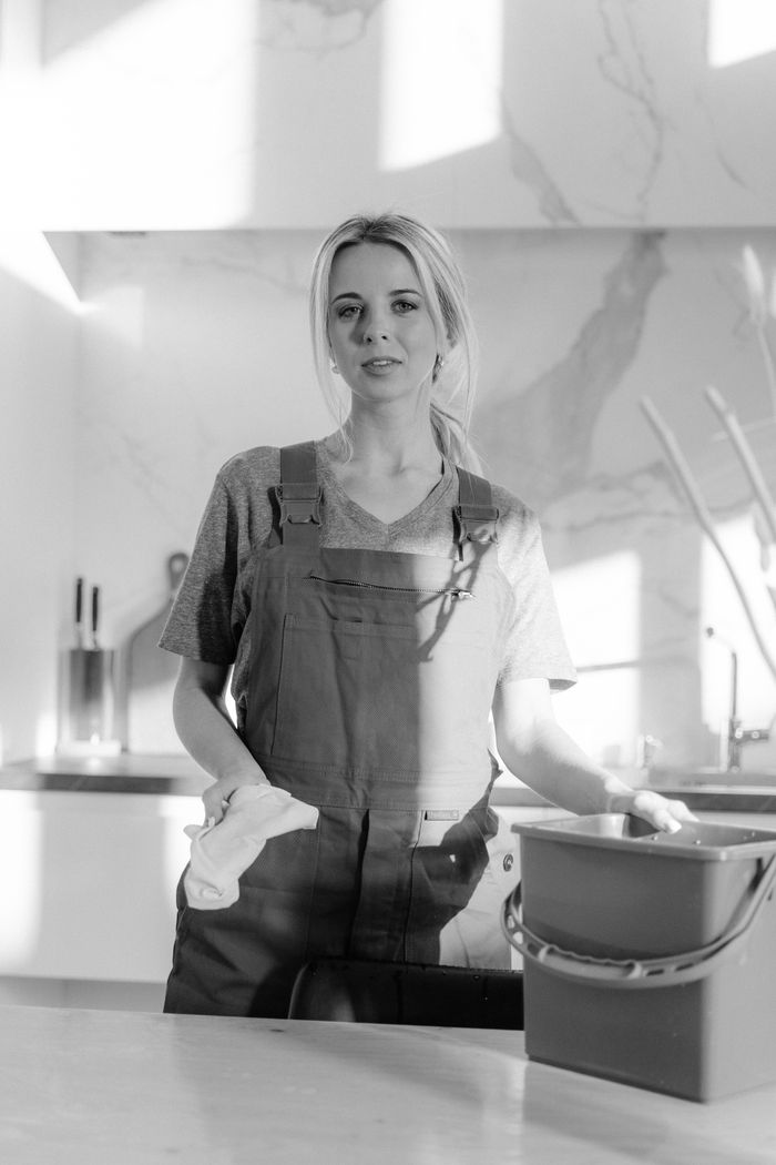 gallery-01 Black and white portrait of a housekeeper with cleaning supplies in a modern kitchen.