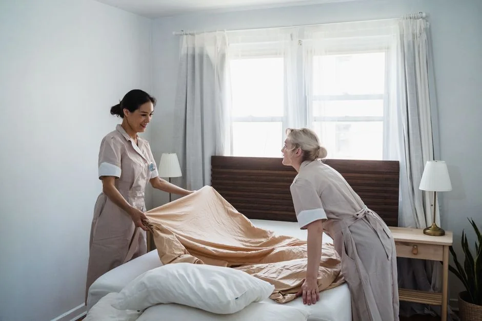 our-story Hotel maid in uniform arranging pillows in a neatly styled guest room.